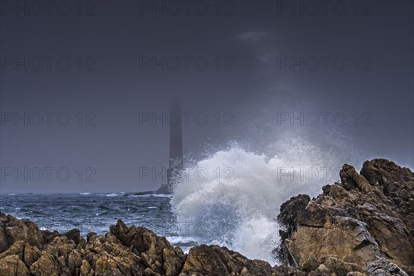 Wave crashing on rock and Phare de Goury lighthouse in the mist at dusk near Auderville