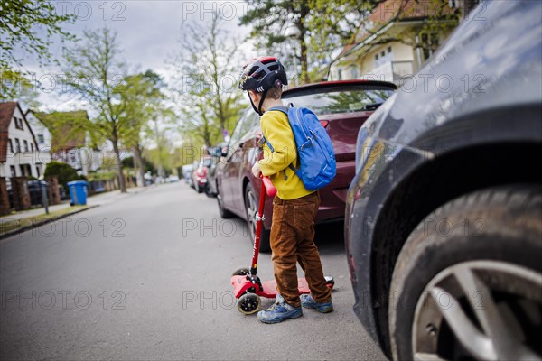 Symbolic photo on the subject of children in road traffic A little boy stands with his scooter between parked cars on a street. Berlin