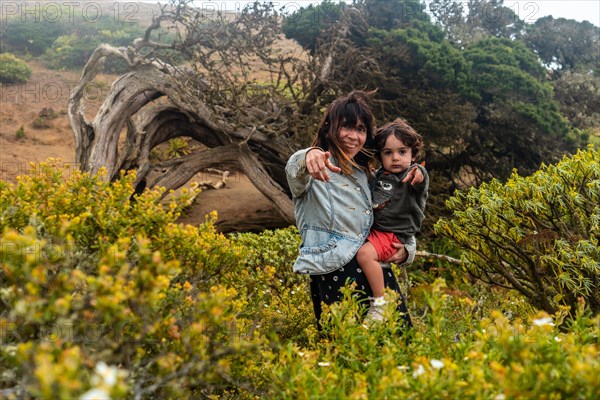 Mother and son looking at the flowers next to a Sabinar tree twisted by the wind of El Hierro and foggy. Canary Islands