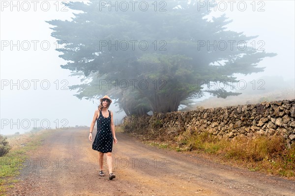 Tourist woman with hat walking through the foggy path towards the juniper forest in El Hierro. Canary Islands