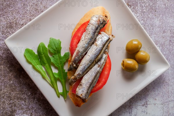 Tapa of sardines on a slice of bread with tomato and olives on a white plate with a typical spanish white background