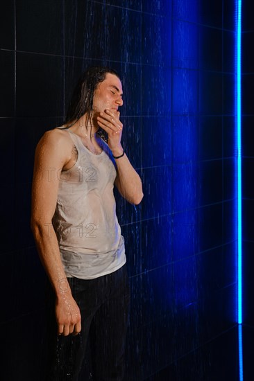 A long-haired man in wet t-shirt stands in a shower in profile illuminated by neon light