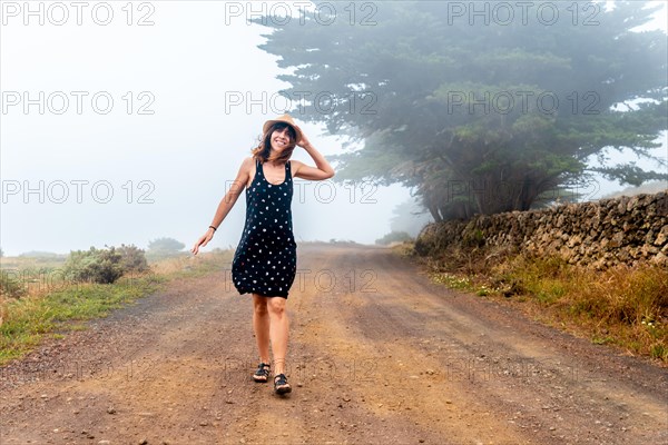 Tourist woman with hat walking through the foggy path towards the juniper forest in El Hierro. Canary Islands