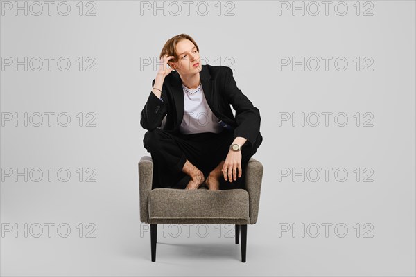 Young barefoot man in black suit sitting in armchair in studio