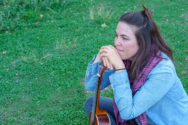 Attractive long-haired brunette girl dressed in blue sitting on the grass looking out of profile holding her ukulele