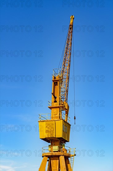 Old and obsolete yellow crane on the harbor pier lit by the afternoon sun with blue sky in the background.