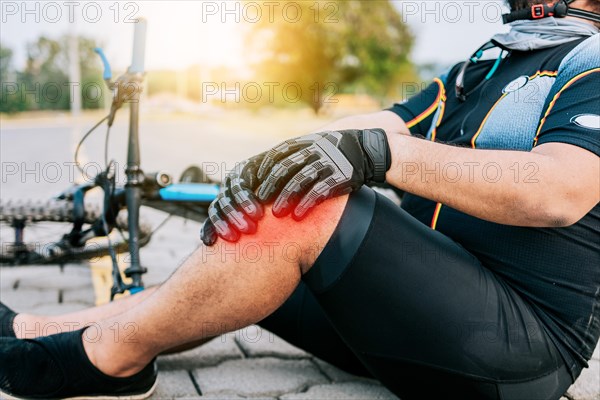 Concept of cyclist with knee injury outdoors. Cyclist with knee pain outdoors. Male cyclist sitting on the pavement with knee pain