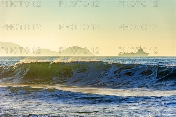 Wave breaking on the beach during sunrise with ship crossing the horizon and mountains in the background.