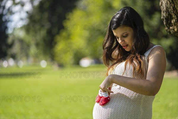 Pregnant latin woman holding baby shoes on her tummy in a park