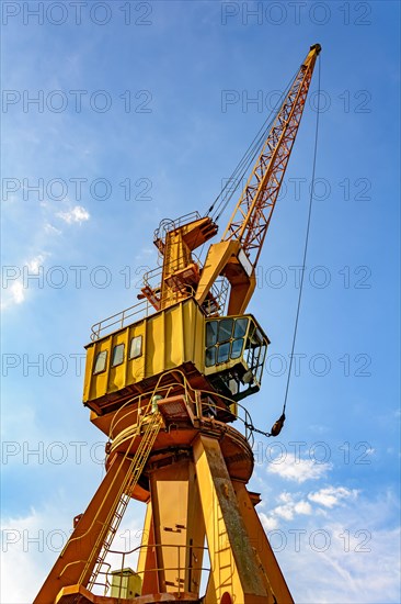 Old and obsolete yellow crane on the harbor pier seen from below and lit by the afternoon sun with blue sky in the background.