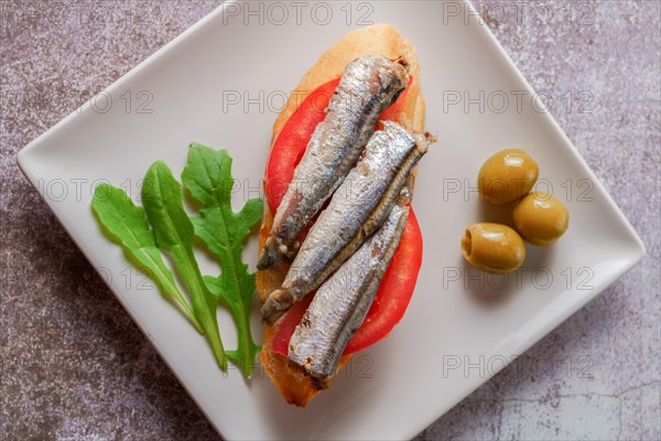 Tapa of sardines on a slice of bread with tomato and olives on a white plate with a typical spanish white background