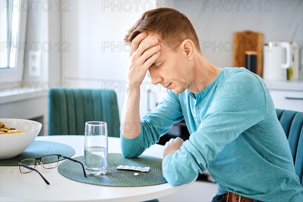 Sick middle aged man in the kitchen in front of pills and a glass of water