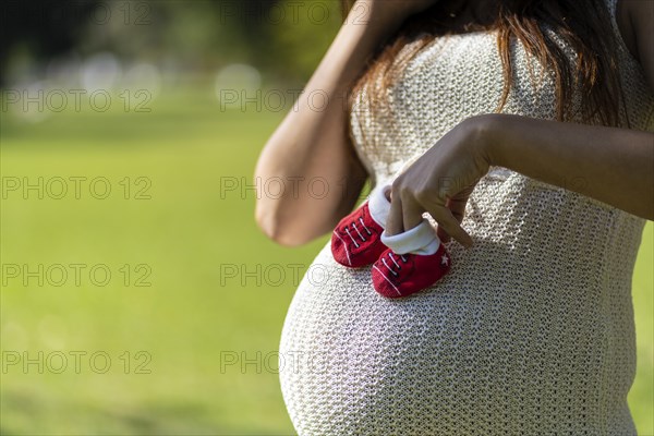 Pregnant latin woman holding baby shoes on her tummy in a park