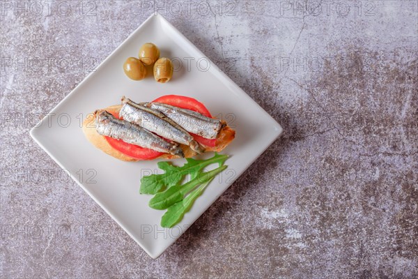 Tapa of sardines on a slice of bread with tomato and olives on a white plate with a typical spanish white background