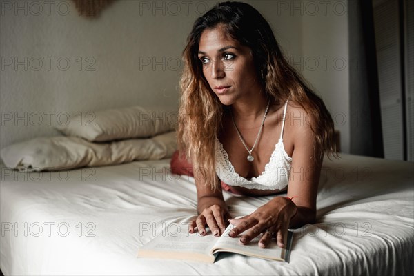 Young latina girl with long hair reading a book lying on her bed is distracted looking towards the window
