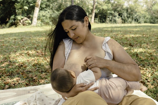 Mother nursing little baby girl while sitting on blanket In a park. Celebrates the act of breastfeeding as an intimate and nurturing moment between the two