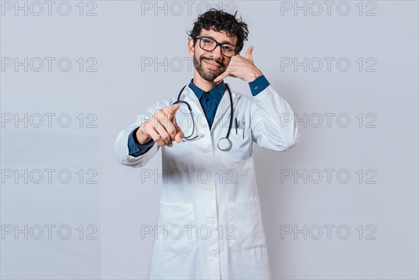 Smiling doctor making call gesture and pointing at the camera. Young doctor making call gesture isolated