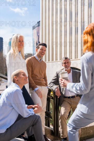 Cheerful group of coworkers outdoors in a corporate office area resting and laughing on a sunny morning