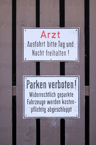 Signs doctor. Exit please keep clear day and night! and Parking prohibited! Illegally parked vehicles will be towed away for a fee mounted on brown painted wooden slats