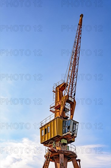 Old and obsolete yellow crane on the harbor pier with blue sky and clouds in the background.