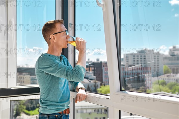 Middle aged man stands on balcony of high-rise building leaning against window frame and drinking juice