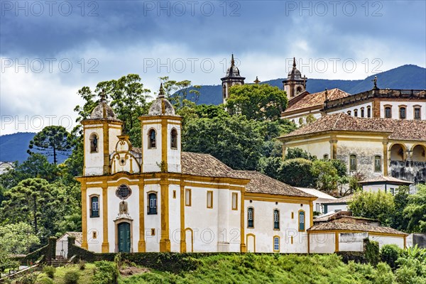 Ancient historical churches among the houses and streets of Ouro Preto city in Minas Gerais with hills and clouds in the background