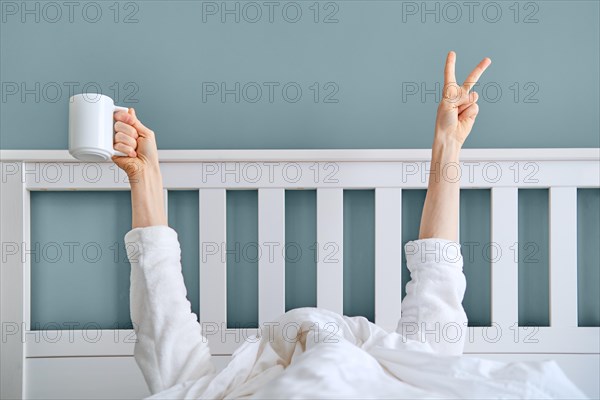 Man hiding under blanket showing hand with victory sign and holding cup of coffee. The early bird catches the worm concept