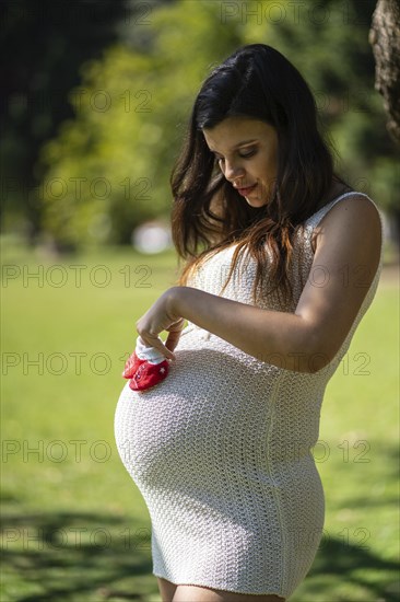 Pregnant latin woman holding baby shoes on her tummy in a park