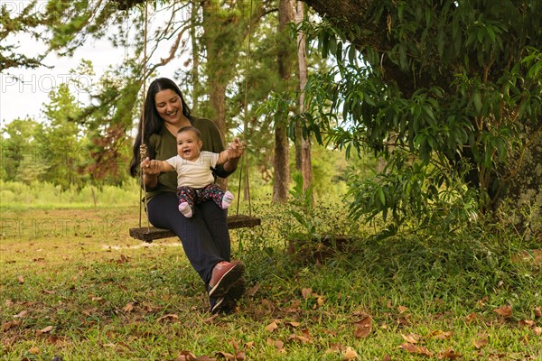 Happy joyful cute baby girl sitting on a swing with her mother playing at the playground