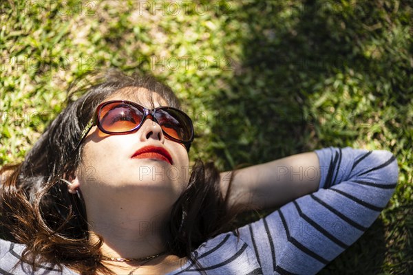 Overhead portrait of an Andean woman with sunglasses lying on the grass with lights and shadows filtered by the tree branches