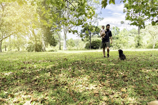 Father holding his baby walking with his dog in the field surrounded by trees. Father's Day Concept