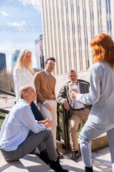 Cheerful group of coworkers outdoors in a corporate office area resting and laughing on a sunny morning