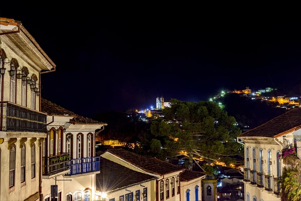 Facades of houses in colonial architecture on an old cobblestone street in the city of Ouro Preto illuminated at night with historic church in background