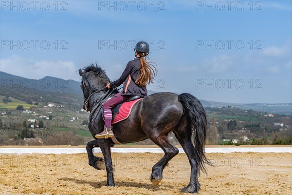 Unrecognizable woman riding on a black horse with a mountainous landscape in the background and blue sky