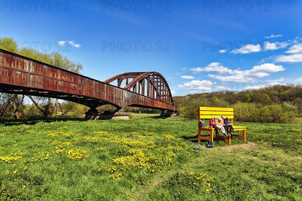 Visitors sitting on large yellow wooden bench in front of old railway bridge