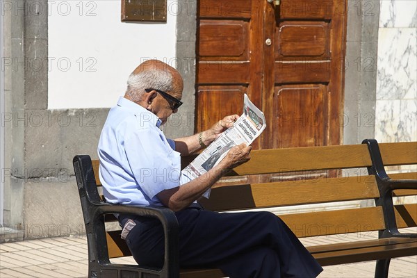 Older man with sunglasses sits on park bench reading newspaper