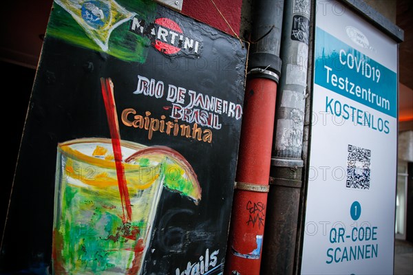 A signboard of a bar advertising Caipirinha cocktails stands next to the sign of a Covid-19 test centre in the old town of Duesseldorf