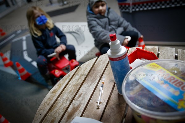 A syringe raised with BioNTech Pfizer children's vaccine lies on a table while children wait in the background at a COVID-19 vaccination and testing centre at Autohaus Olsen in Iserlohn