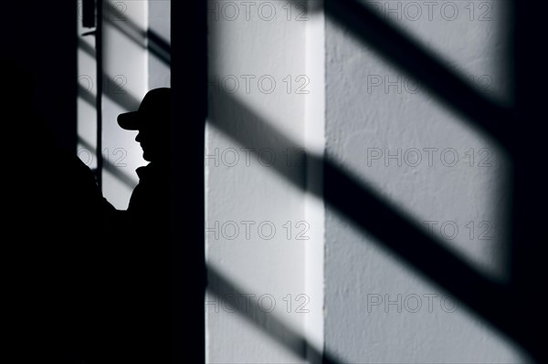 Symbolic photo on the subject of male criminality. The silhouette of a man wearing a baseball cap stands out between shadows of a window.Berlin
