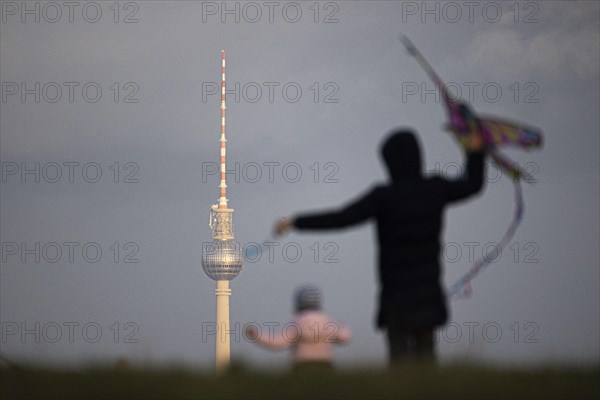 An adult and a child stand out while flying a kite with a view of the Berlin TV Tower in Berlin