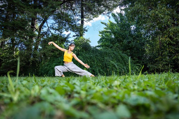 Woman doing qi gong tai chi exercise. A woman practices tai chi in nature surrounded by trees and a blue sky. Close-up of the grass