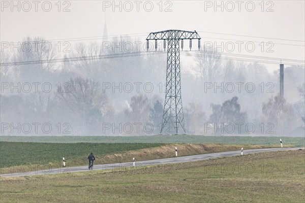 A cyclist rides on a country road in front of an electricity pylon