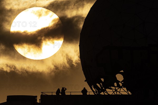 The silhouettes of people and the former listening station on Teufelsberg stand out against the setting sun in Berlin