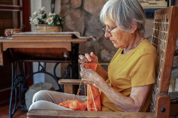 Profile portrait of an old woman sitting in an armchair at home knitting with 2 needles