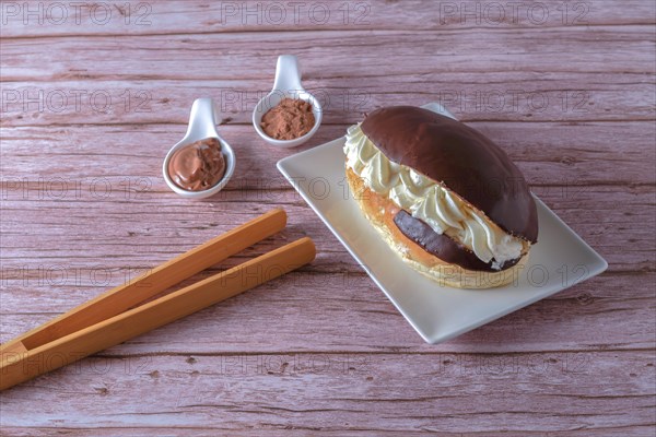 Delicious chocolate-covered cream eclair and white ceramic spoons with chocolate and cocoa on the side with wooden tongs