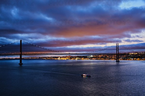 View of 25 de Abril Bridge famous tourist landmark of Lisbon connecting Lisboa and Almada on Setubal Peninsula over Tagus river in the evening twilight with tourist boats. Lisbon