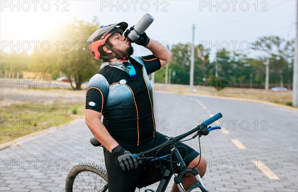 Tired cyclist drinking water outdoors. Chubby cyclist on his bike drinking water