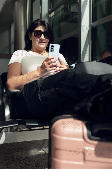 Passenger female traveler at airport waiting for air travel using smart phone. Mature business woman smiling sitting leaning her legs on travel suitcase