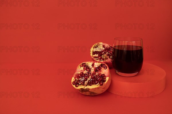 Glass of pomegranate juice on a podium with a pomegranate divided into pieces. Red bright background. Close up. Place for text. Healthy lifestyle concept