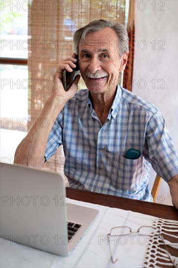 Mature business man smiling and talking on phone in front of laptop looking towards camera